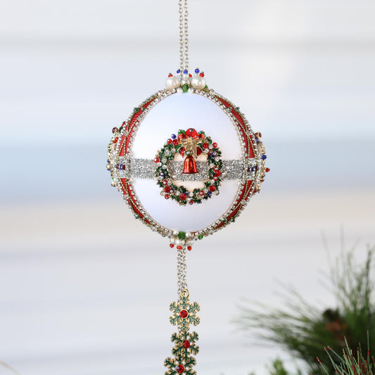 Handmade satin beaded white Christmas ornament featuring metal alloy wreath earring, tassel with snowflakes, and trim, hanging in front of gray background and holiday greenery by Whim5seasons.