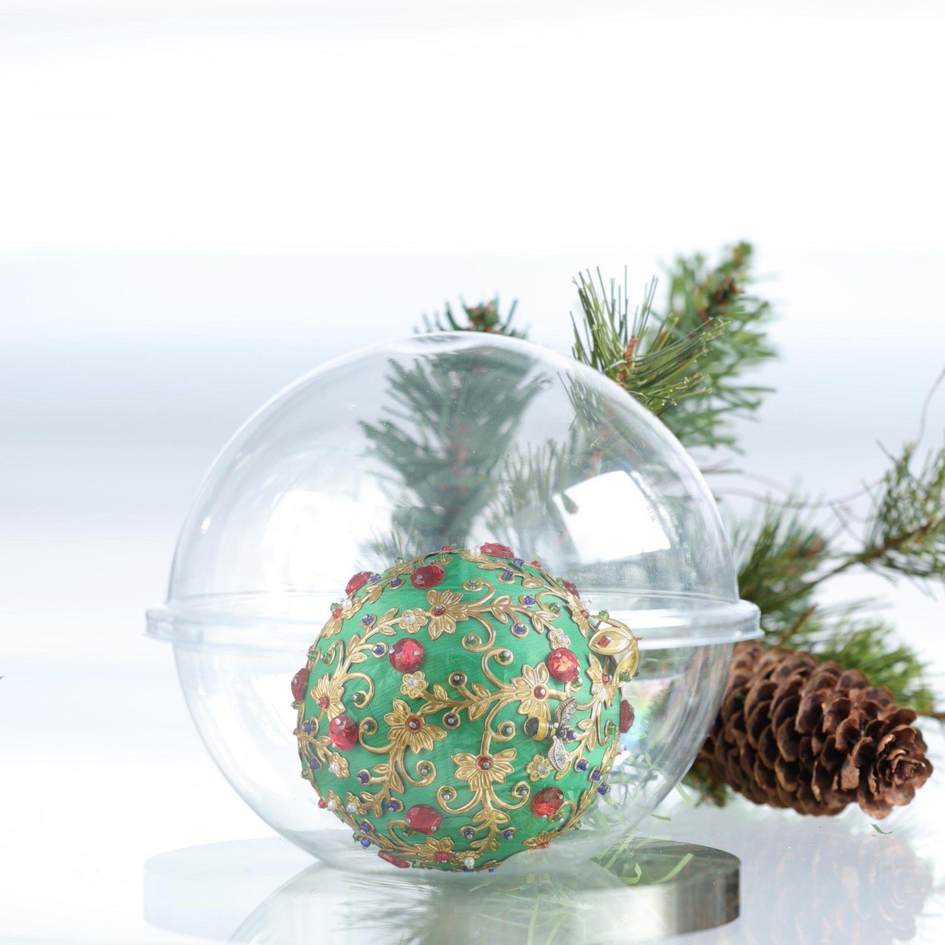 Decorative handmade Christmas satin ball with gold and red details inside a clear glass storage container, surrounded by pine branches and a pine cone by Whim5seasons.
