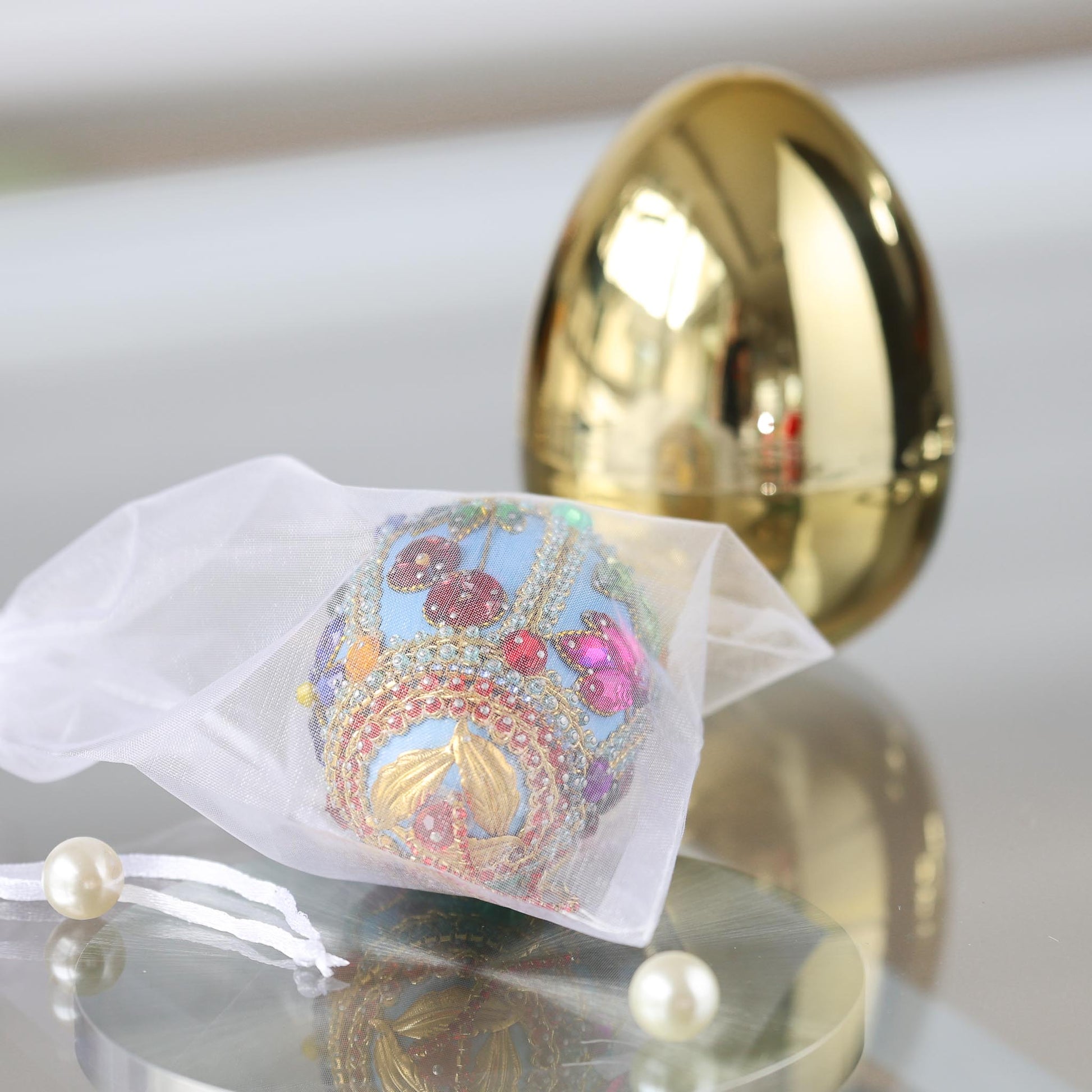 Handmade Easter push pin ornament Faberge style in organza bag with a large gold egg container in the background on a reflective surface by Whimseasons.