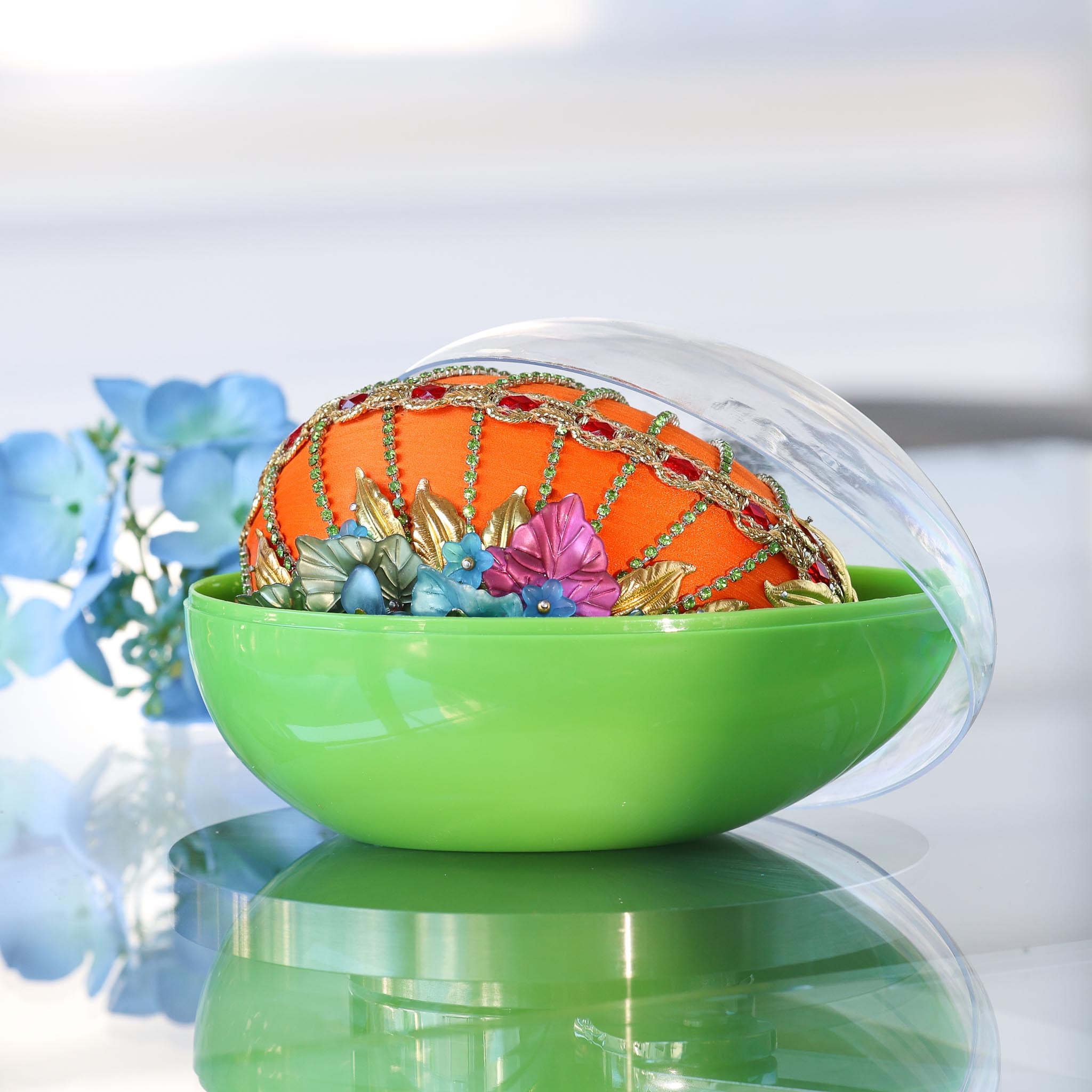 Handmade Easter Egg - a completed push pin ornament, orange with red rhinestones and colorful flowers inside a green storage container on a reflective surface.
