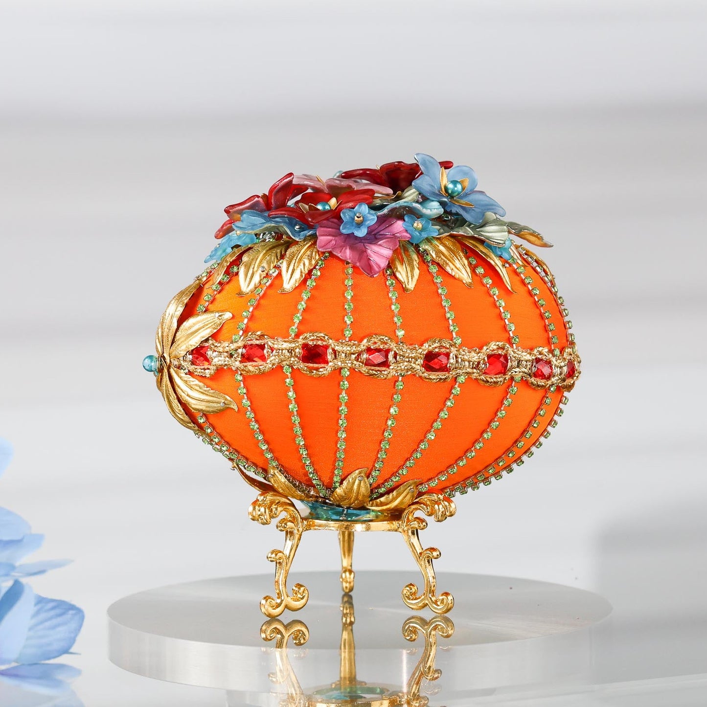 A side detail of a handmade orange Easter egg satin ornament with embellishments including red rhinestones, gold trim, and beads. The ornament is displayed on a gold stand by Whim5seasons.