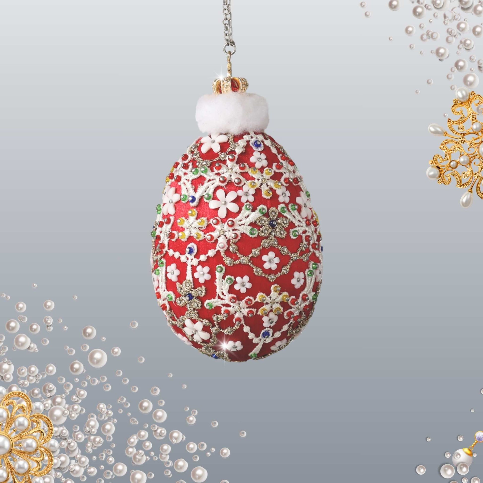 A decorative hanging ornament shaped like an egg, featuring a red background covered with white floral patterns and multicolored jewel-like embellishments. It has a fluffy white top and is suspended by a chain against a light gray gradient background with pearl and gold decorative corner accents.