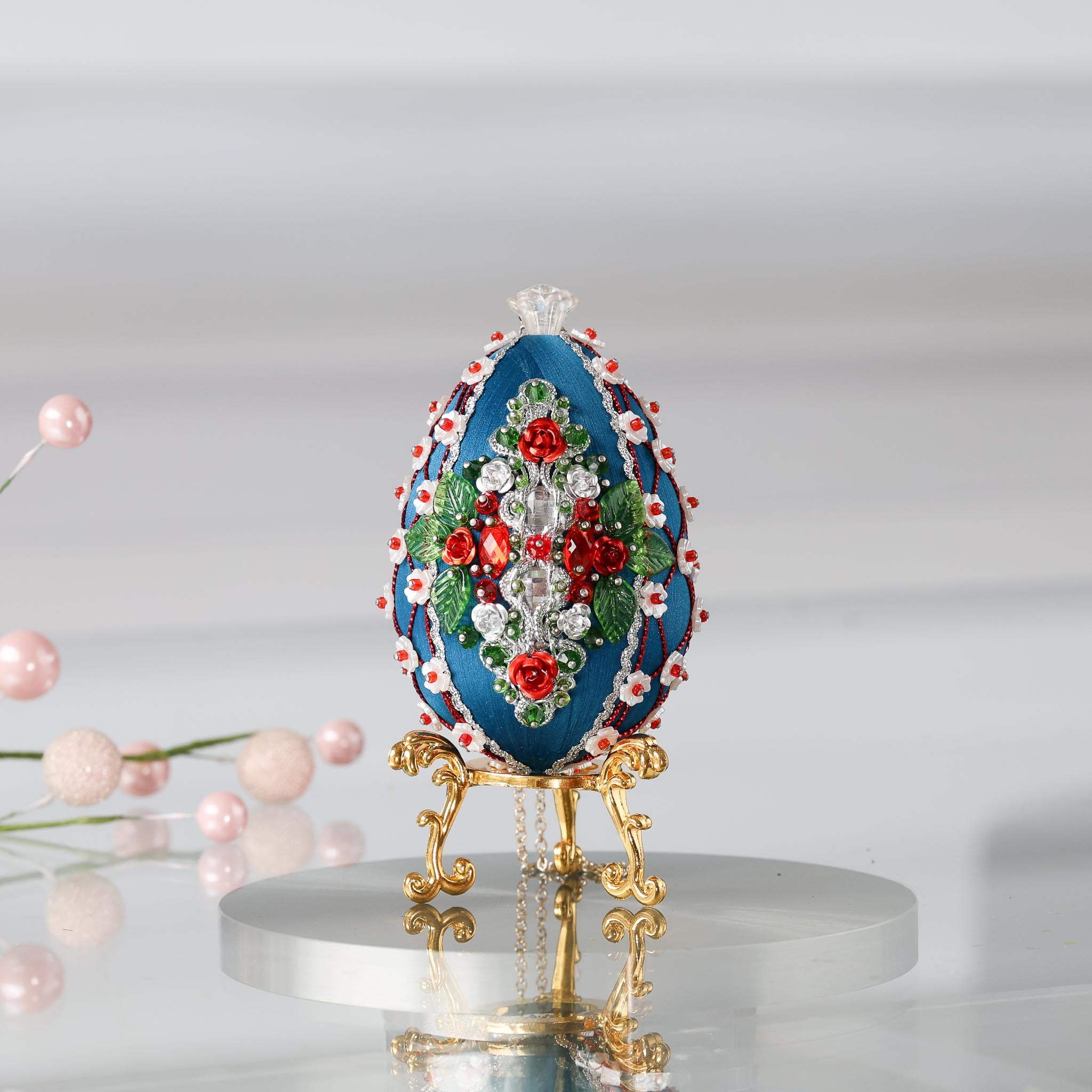 Luxurious handmade dark teal push pin Easter Egg ornament on a gold stand set against a light background featuring red roses, silver cording, and sparkly beads.  (Dark Slate Blue)