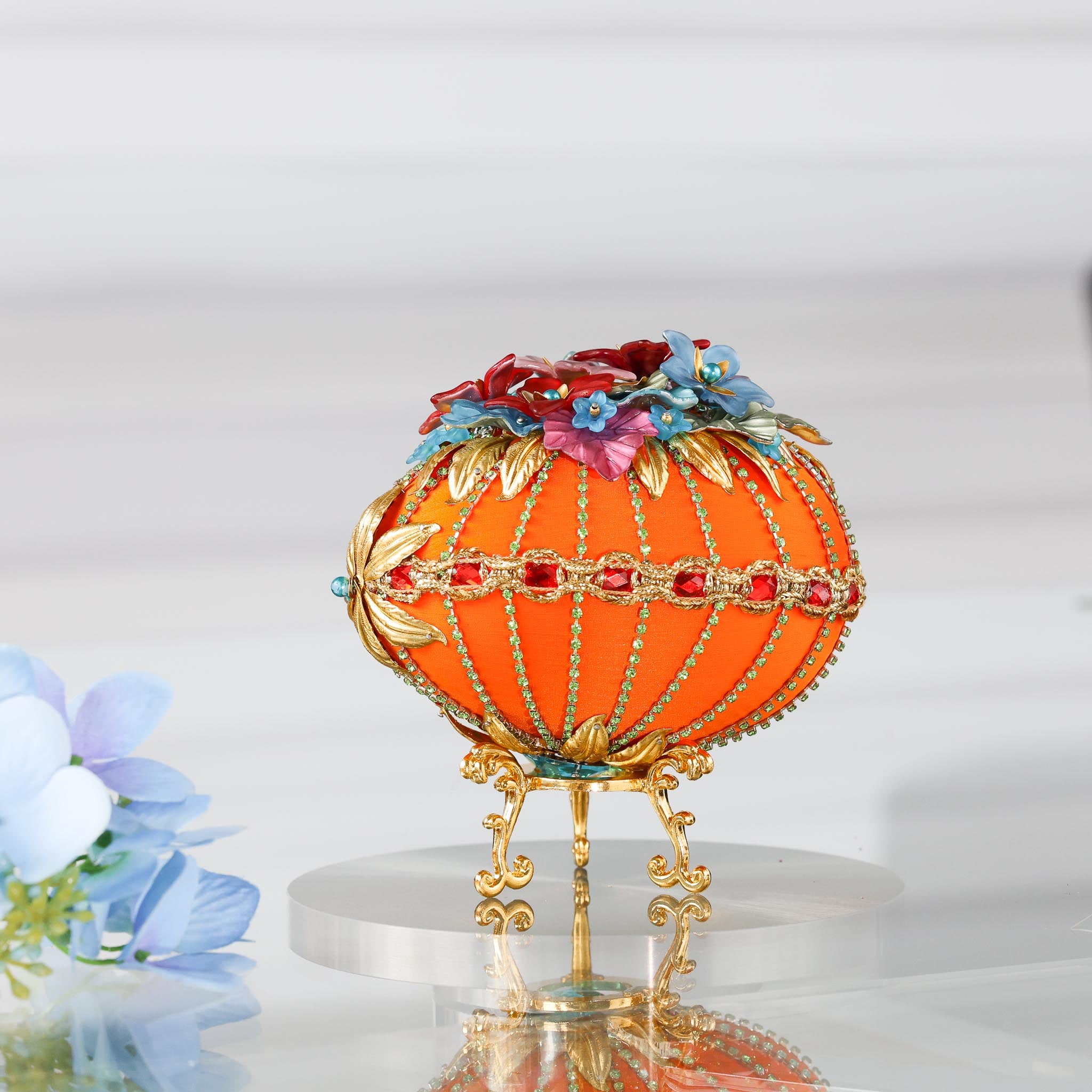 A side detail of a handmade orange Easter egg satin ornament with embellishments including red rhinestones, gold trim, and beads. The ornament is displayed on a gold stand.