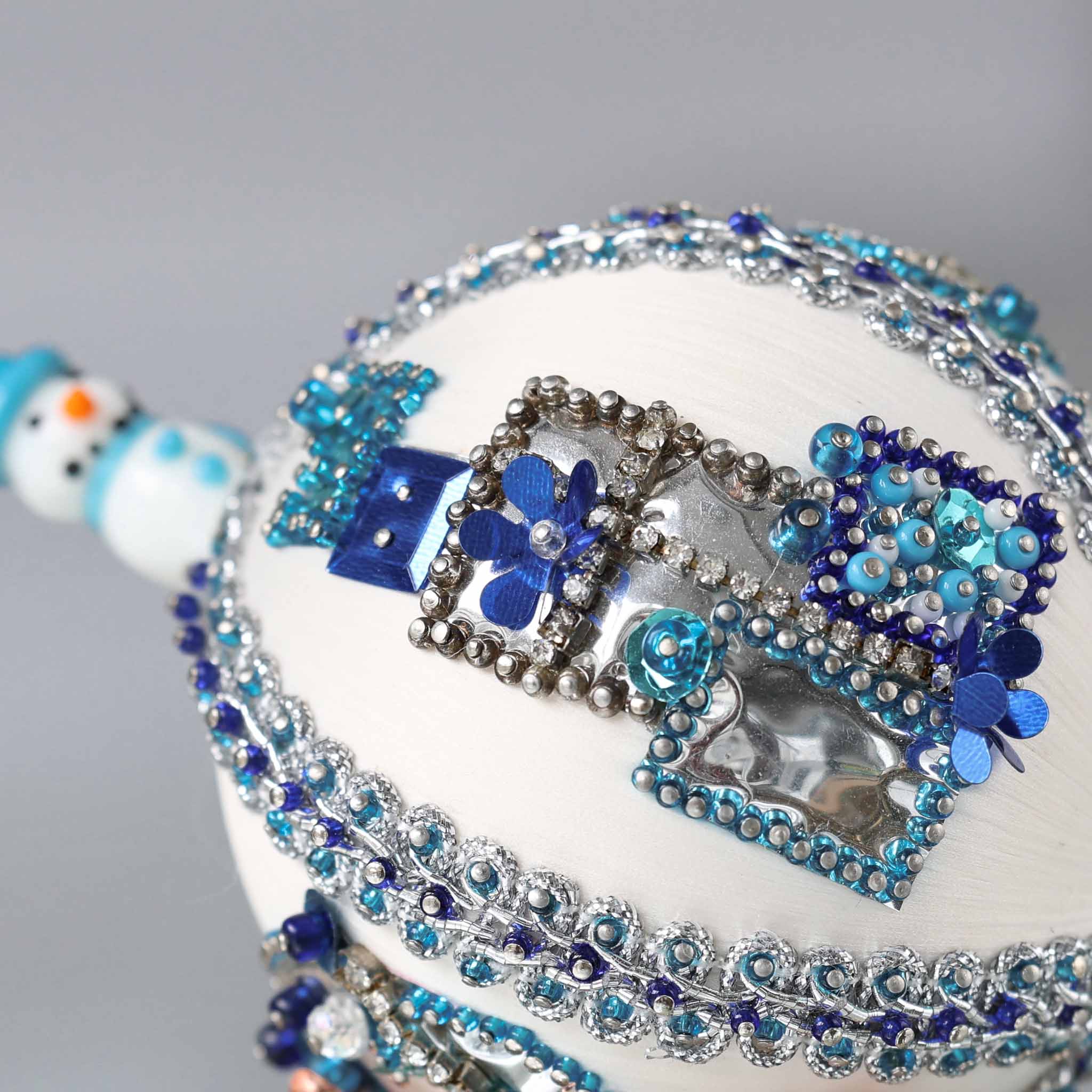 Close up of white handmade Christmas beaded push pin ornament in with sequined blue gift boxes and a snowman porcelain bead in the background.