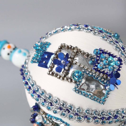 Close up of white handmade Christmas beaded push pin ornament in with sequined blue gift boxes and a snowman porcelain bead in the background.