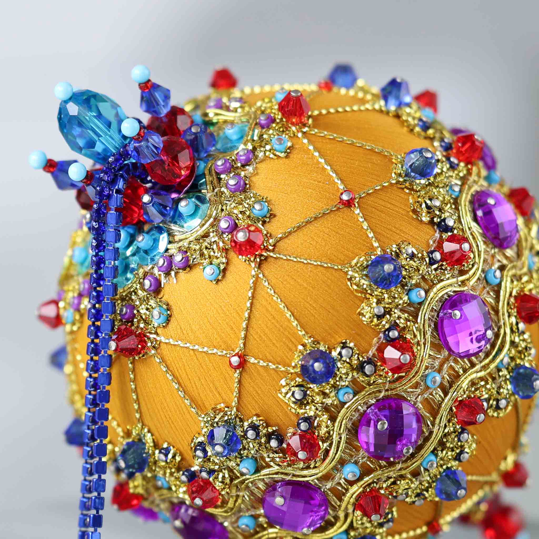 A close-up picture of a handmade Christmas push pin ornament showing a top of an ornament embellished with dark blue rhinestone chain, blue sequins, and red and blue glass beads. (Gold)