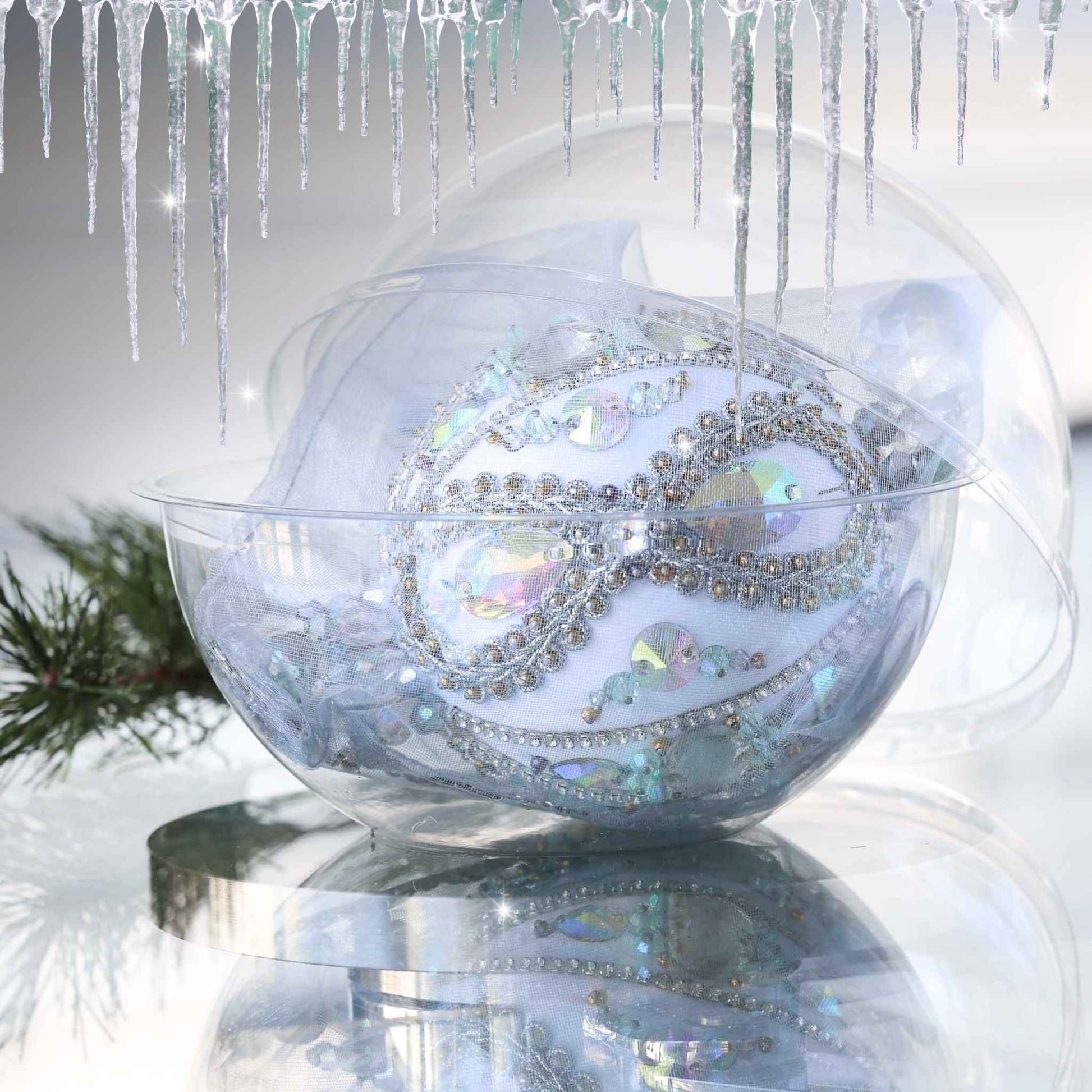 Handmade Christmas beaded ornament with intricate design on a reflective surface in a clear storage box with an evergreen branch in the background.