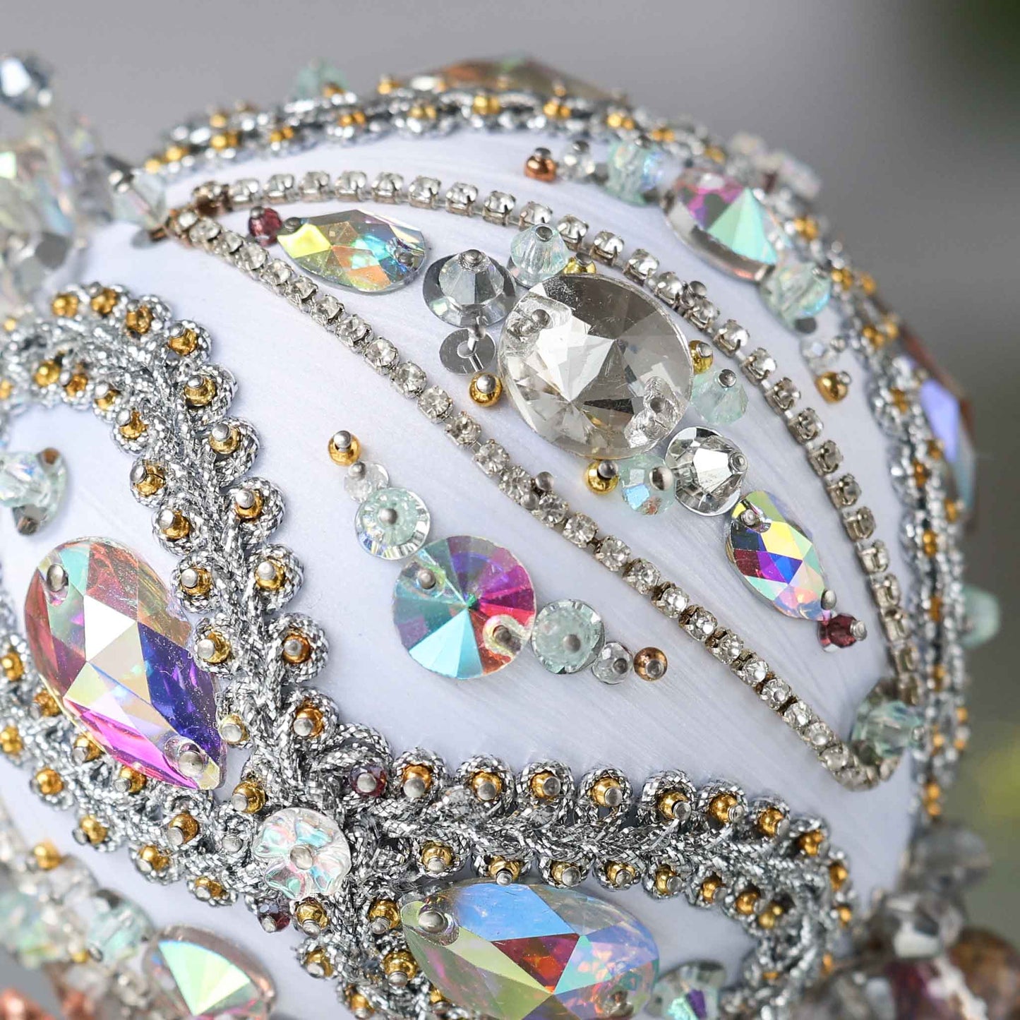 Close up of a handmade white Christmas beaded ornament featuring large glass AB rhinestones, silver trim, and hundreds of seed beads, pinned to a satin core with dressmaker pins.