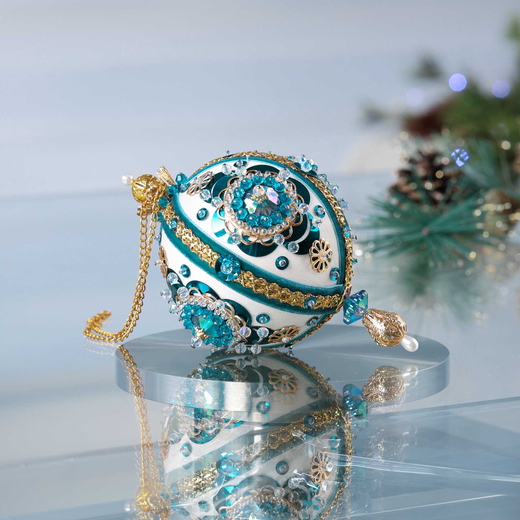 Handmade 1970 retro mid century white satin push pin ornament featuring large AB rhinestones, blue crescent sequins, and a filigree gold bead on the bottom with a Christmas branch in the background.
