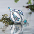 Load image into Gallery viewer, Handmade Christmas satin push pin ornament in white with blue and silver sequined tree, snowman bead, and a large Tibetan style bead on the bottom on a reflective surface.
