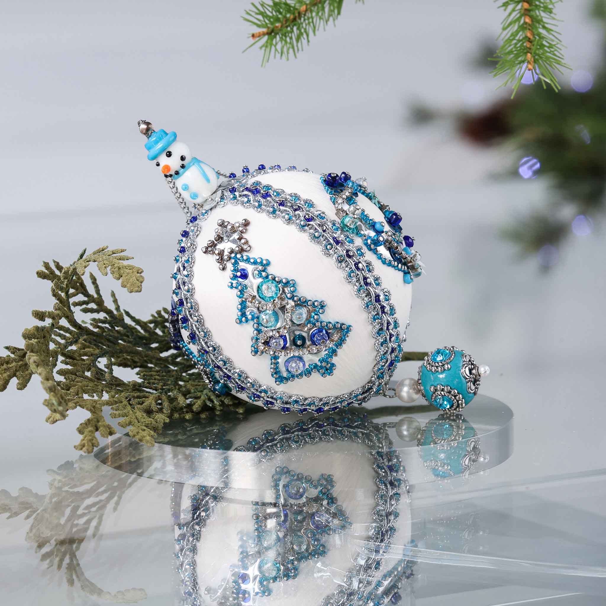 Handmade Christmas satin push pin ornament in white with blue and silver sequined tree, snowman bead, and a large Tibetan style bead on the bottom on a reflective surface.