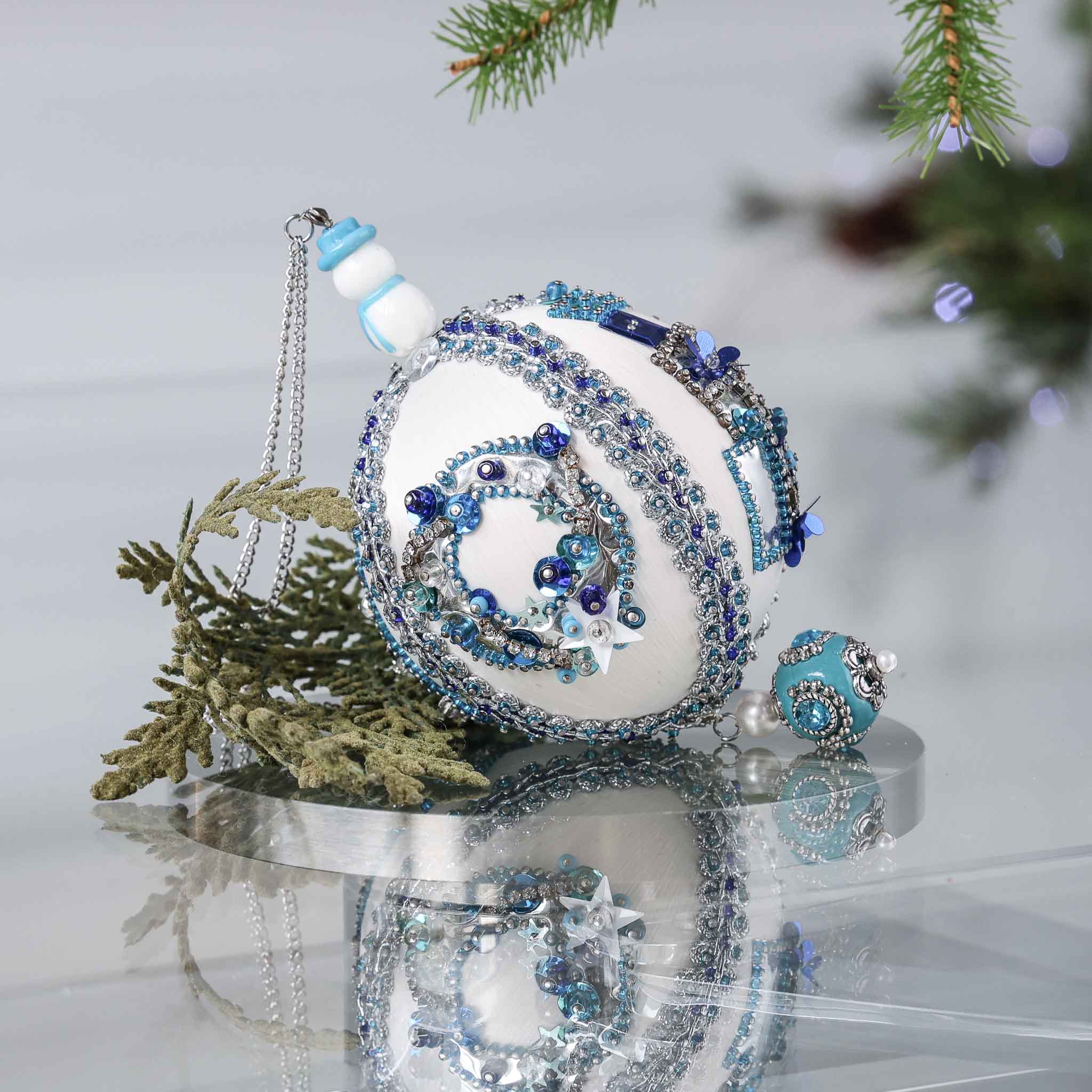 Handmade Christmas satin push pin ornament in white with blue and silver sequined holiday wreath, snowman bead, and a large Tibetan style bead with green branches on a gray background.