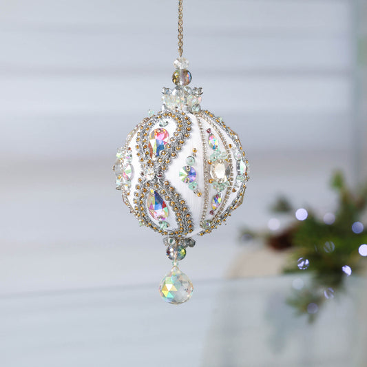 Handmade white satin push pin ornament beaded with glass AB rhinestones, and hundreds of tiny seed beads, hanging on a chain in front of light gray background.