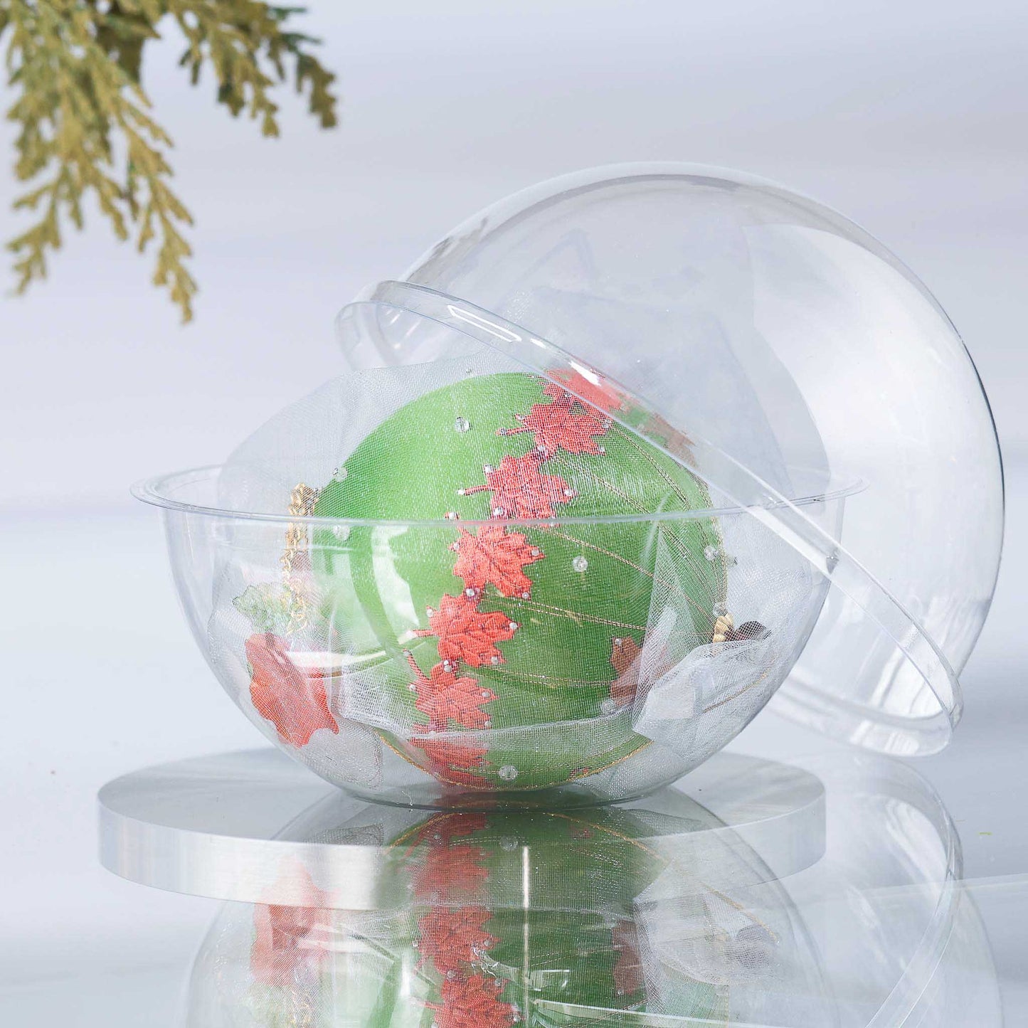 Completed Push Pin Ornament "Leaves Are Falling" features an olive colored satin foam core and comes in a gauze bag and a clear plastic storage container, pictured on top of the reflective surface. (Olive)