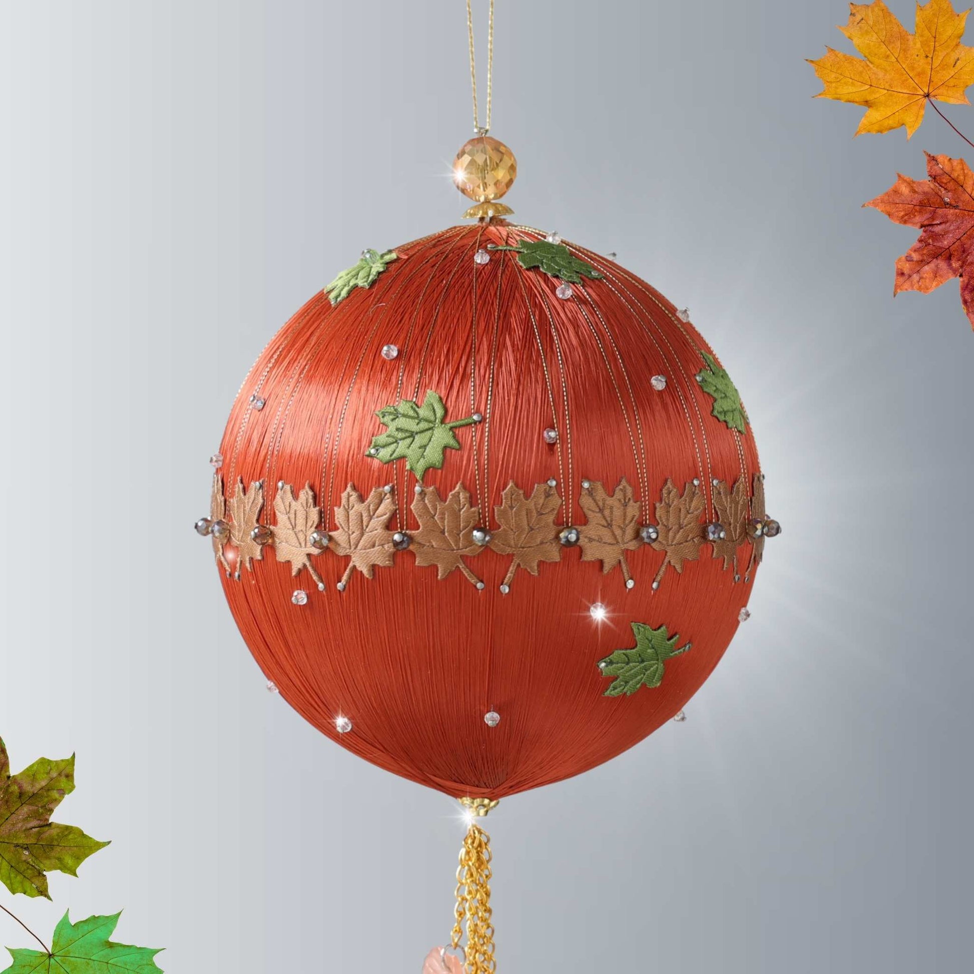 A handmade satin beaded and sequined  ornament decorated with metallic gold, maple leaves, small green leaves, suspended against a light gray background with autumn leaves in the corners by whim5seasons.