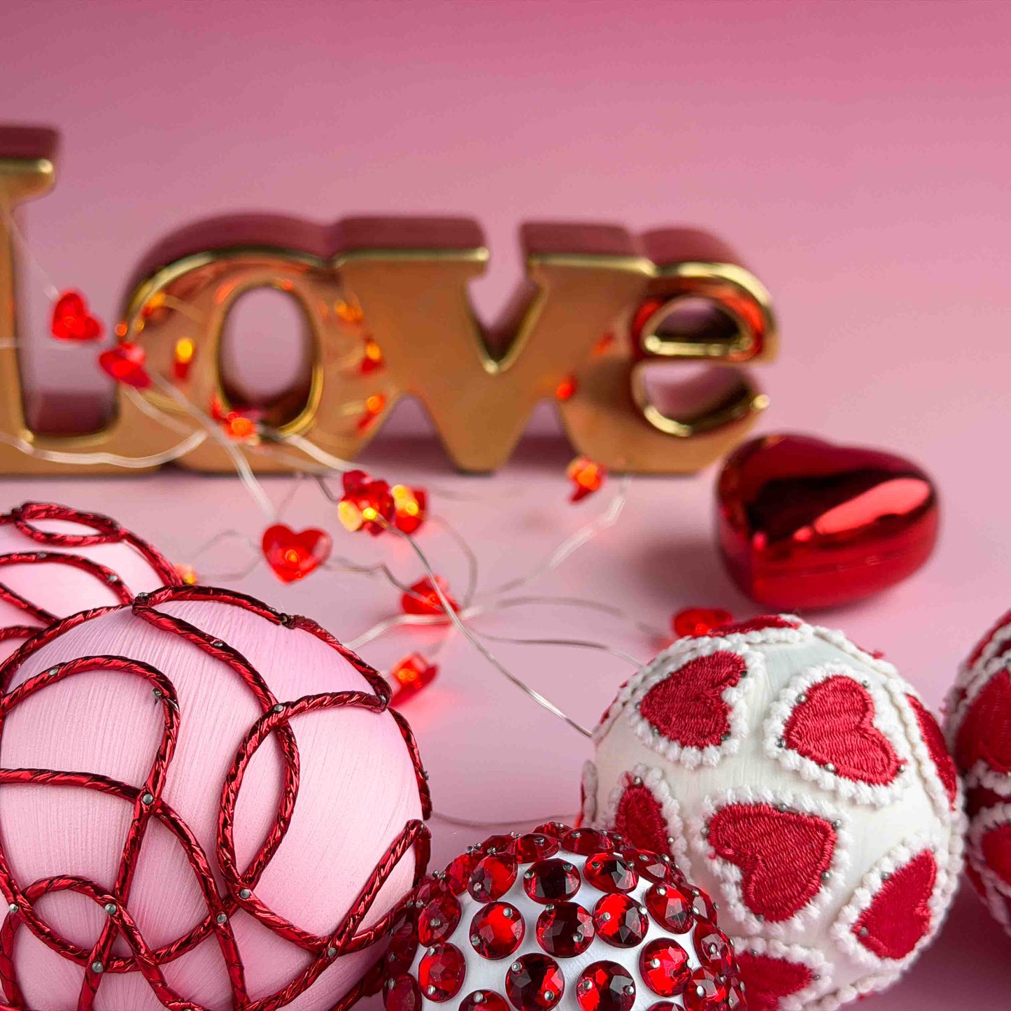 Close up of satin handmade filler beaded ornaments showing rhinestones and heart trim pinned to foam balls with dressmaker pins by Whim5seasons, with 'Love' spelled out in gold letters on a pink background.