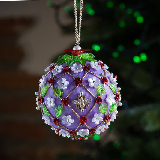 Handmade Christmas ornament with floral patterns and charms, hanging against a blurred green background with green Christmas lights. (Medium Purple).