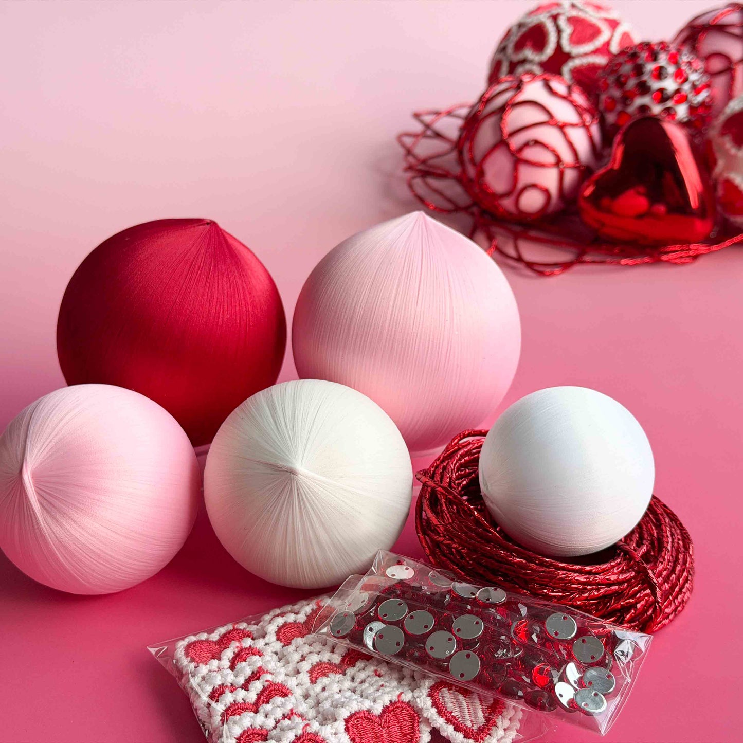 Materials for a beaded satin ornament kit project - decorative items including red, pink, and white foam core balls, heart trim, and red rhinestones on a pink background by Whim5seasons